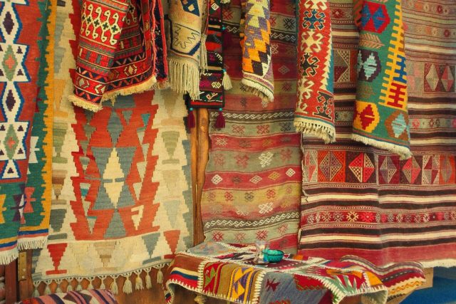 Vibrant handmade rugs with geometric patterns hung in a traditional market setting.